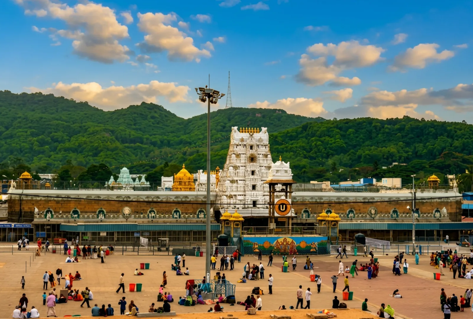 A wide horizontal landscape shot showing the grand white tiered tower of the Tirumala Venkateswara Temple complex situated against lush green hills under a cloudy sky.