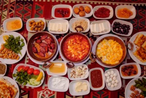 A high-angle view of a table filled with various copper and white bowls containing eggs, cheeses, olives, sliced tomatoes, cucumbers, honey, and sausages.