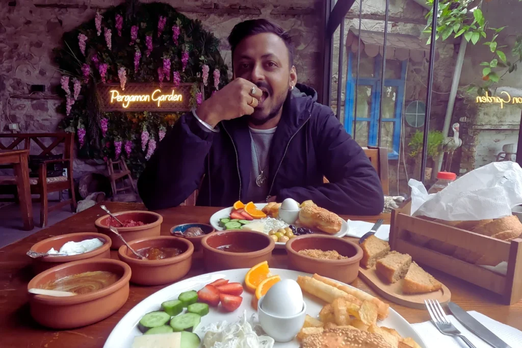 Turkish Breakfast 2 Akash sitting at a wooden table outdoors, smiling while enjoying a comprehensive Turkish breakfast spread served in small copper bowls.