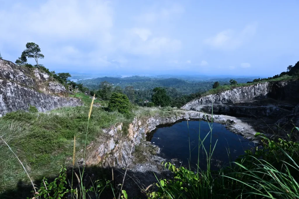 MANJAPPARA QUERRY 1 Rocky quarry cliffs opening into a valley with a small pond visible amidst lush green hill country in early morning light.