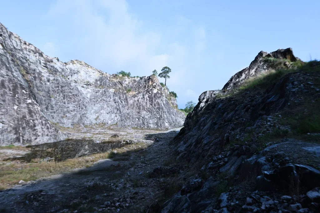 MANJAPPARA QUERRY 2 A wide view inside a steep stone quarry canyon with rocky cliffs towering on either side under a bright blue sky.