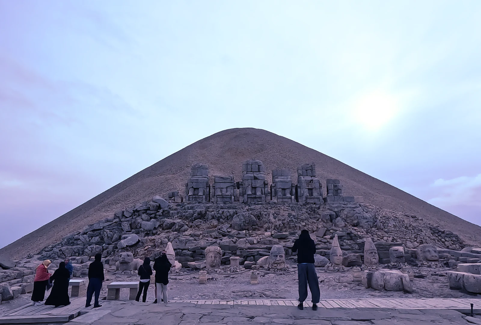 A wide view of the ancient, massive stone head sculptures scattered in front of the elevated stone tumulus peak of Mount Nemrut under a clear sky.