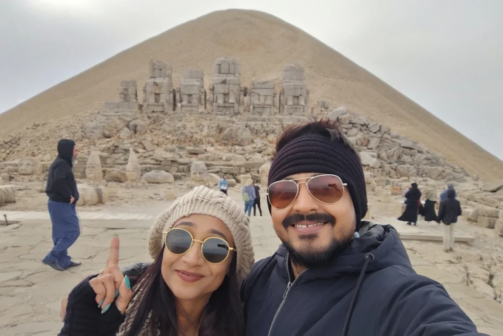 MT. NEMRUT - 6 A selfie of Akash & Sumana smiling outdoors in front of the giant stone head statues and tumulus peak of Mount Nemrut.