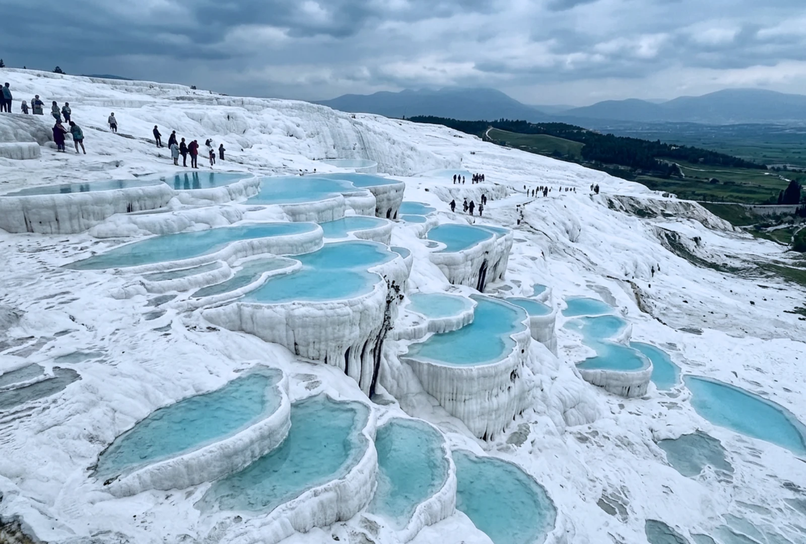 An aerial view of white calcium travertine terraces holding vibrant blue thermal water under a clear sky.
