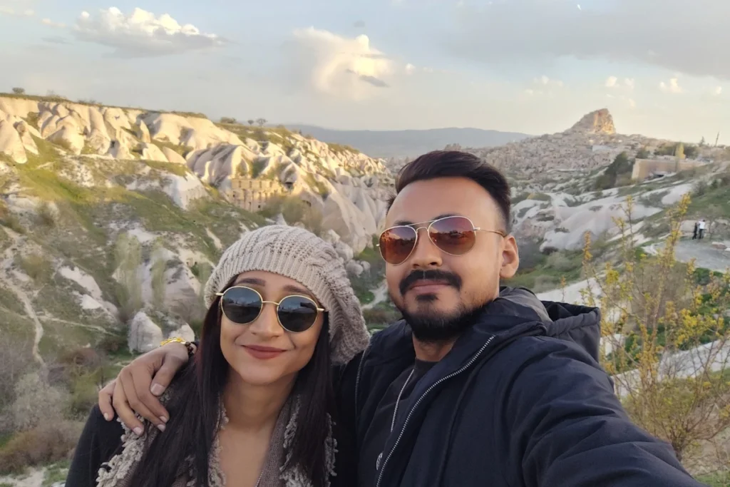 Pigeon Valley 1 A selfie of Akash & Sumana smiling outdoors with the sprawling valley and cliffside rock formations behind them.