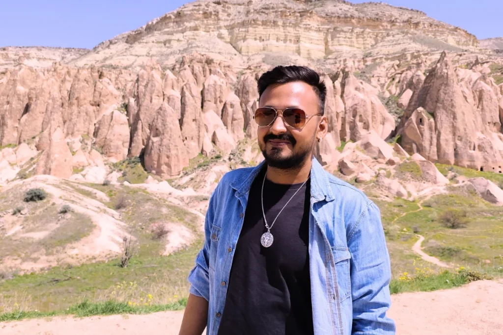 Red Valley 2 Akash wearing sunglasses, a denim jacket, and a beanie smiling in front of the red rock canyon backdrop.