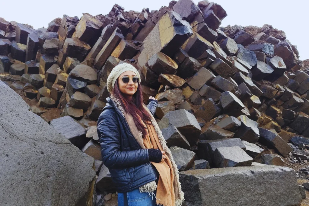 Siverek Basalt Columns 1 Sumana in a dark jacket, beanie, and sunglasses sitting atop a pile of ancient hexagonal basalt rock columns outdoors.