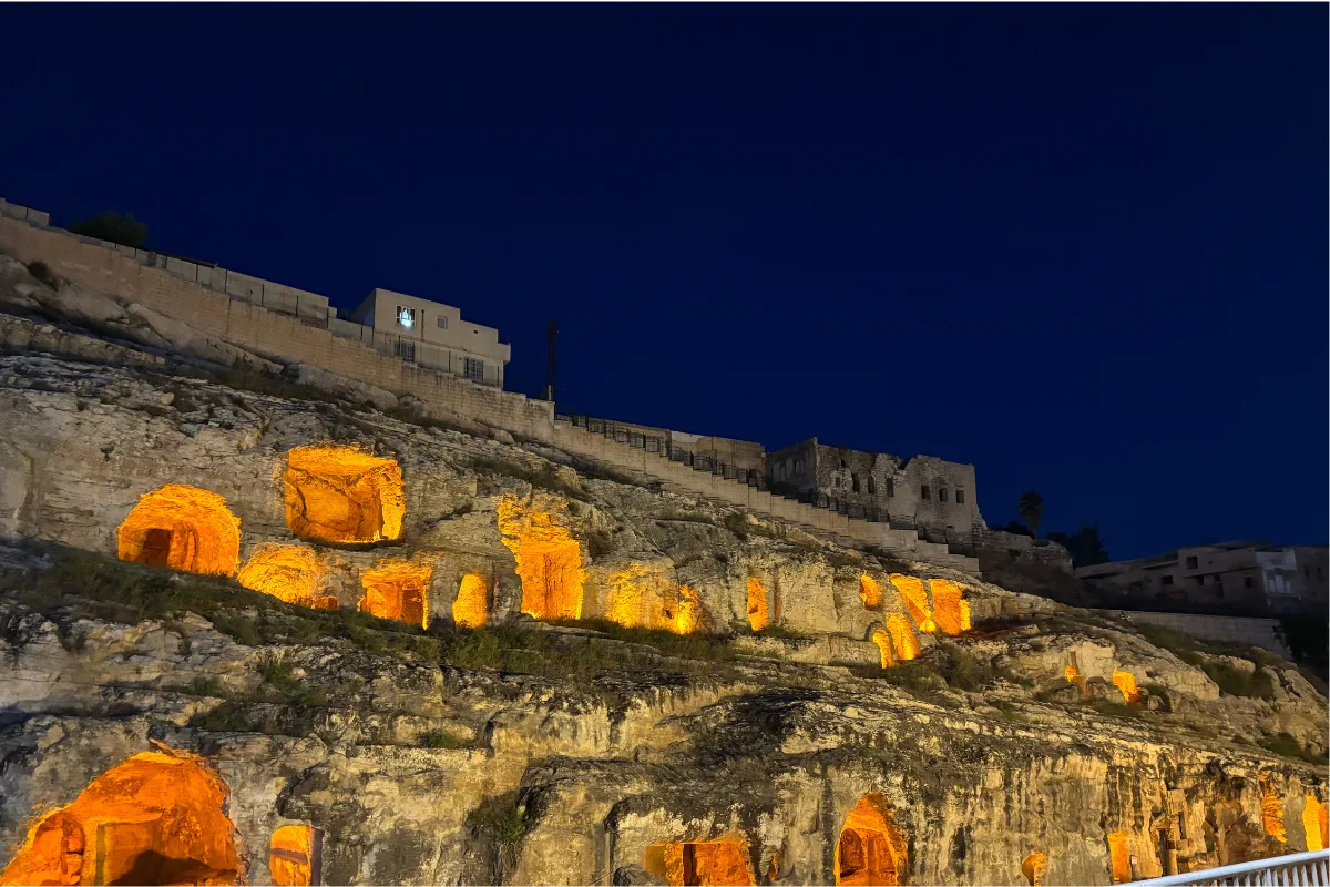 Wide panoramic night view of the illuminated Kızılkoyun Necropolis, showing ancient rock-cut tomb openings glowing with warm yellow lights set into a stone hillside in Şanlıurfa.