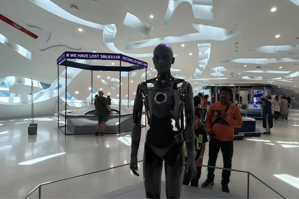 Museum of the future 1 Futuristic realistic humanoid robot named Ameca standing on display in a dark exhibit hall surrounded by visitors.