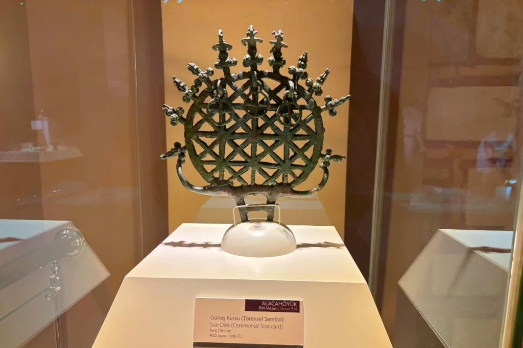 Alacahoyuk 1 An intricate openwork bronze ceremonial sun disk artifact featuring geometric patterns and animal motifs on a display stand.