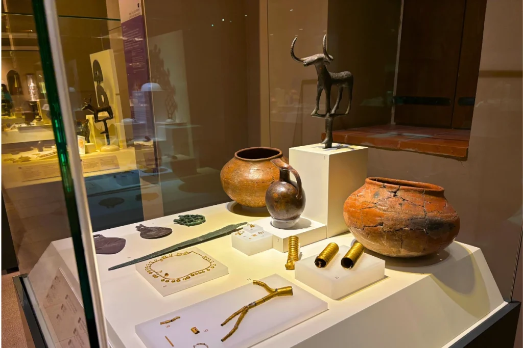 Alacahoyuk 4 Collection of ancient Bronze Age artifacts including a bull statue, pottery, pitcher, and jewelry arranged inside a display case.