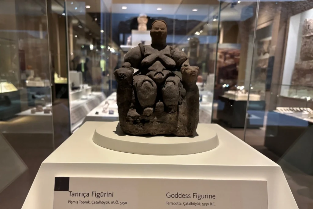 CatalHoyuk 13 Small ancient terracotta figurine of the Mother Goddess displayed on a pedestal inside a glass museum case.