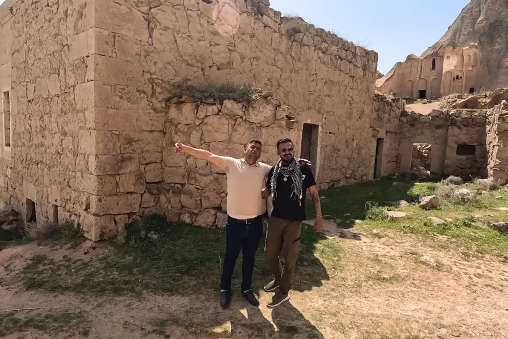 Selime Monastery8 Two men standing together near an ancient stone wall structure, with one man pointing toward the architectural details.
